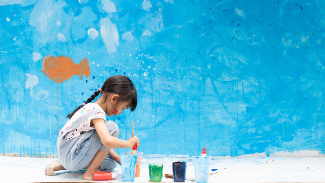 Adorable 5 years old asian little girl is smiling while painting the wall with water color at home, concept of art education for kid, homeschooling and learning activity by playing for child.