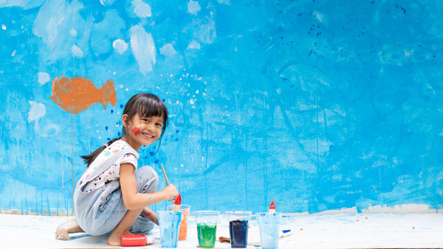 Adorable 5 years old asian little girl is smiling while painting the wall with water color at home, concept of art education for kid, homeschooling and learning activity by playing for child.
