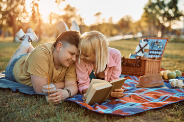 Affectionate female couple enjoys in reading book on picnic in nature.