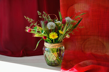 Still life with a bouquet of wild flowers and herbs in a glass vase against a background of red fabric.