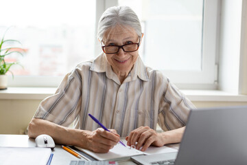 Friendly smiling caucasian senior woman in eyeglasses sits at the laptop with a pen in hand at home, selective focus.