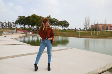 Young and beautiful red-haired woman listening to music with headphones in a park where there is a large lake. The woman is happy and dancing. Concept expressions and happiness, music and dancing.