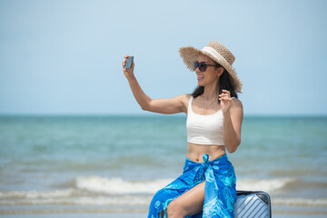 Cheerful young woman at beach with hat, sunglasses, suitcase. Happy smiling girl enjoying  taking a selfie the beach. asian tanned woman feeling refreshed in yellow scarf during summer vacation