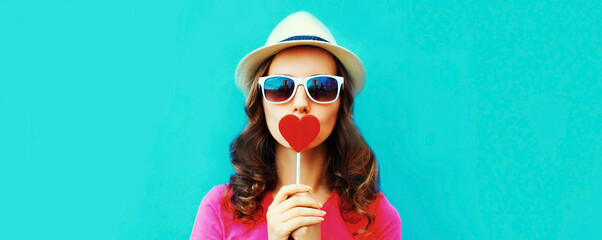 Portrait of young woman with red heart shaped lollipop wearing summer straw hat on blue background