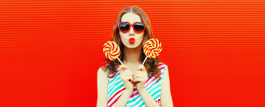 Portrait of happy young woman with colorful lollipop blowing her lips wearing heart shaped sunglasses on red background, blank copy space for advertising text