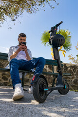 Young man looks at smartphone on bench next to electric scooter
