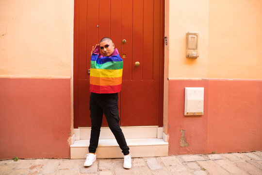 Non-binary person, of South American origin and young, the person is wearing make-up and holding the gay pride flag on a red background. Concept equality, homosexuality, gay, transgender, gay pride.