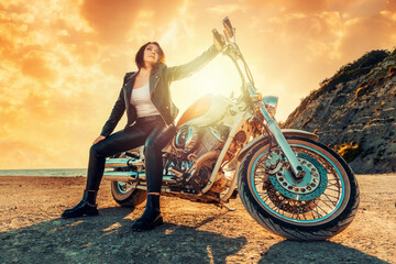 Fototapeta na wymiar A beautiful cool woman in a leather jacket and pants, posing sitting on a motorcycle, against the background of an epic sunset. Bottom view. The concept of motorcycle travel