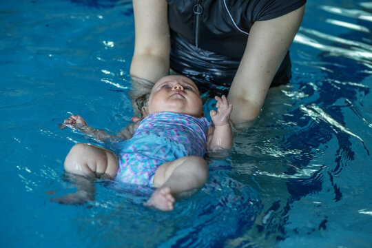 Mom supports baby with hands in swimming pool, teaches to swim and dive.
