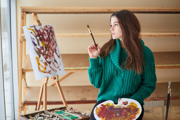 brunette artist girl stands near the easel and hoslt with brushes in her hands.