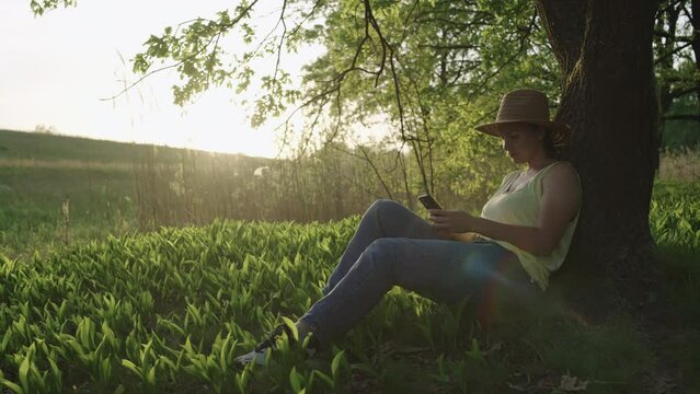 Caucasian woman sitting on the grass in the park with a phone. The girl works in business, orders food, book a hotel through a mobile device in a landscape place in nature.