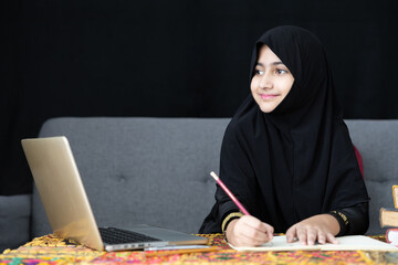 muslim girl writing on notebook, using a laptop computer and online learning at home