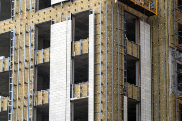 Monolithic concrete frame of apartment building under construction with partially attached facade cladding blocks. Energy efficiency. Installation of external wall thermal insulation with rock wool.