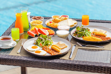 Hotel breakfast. Pair having breakfast on tropical villa near swimming pool. Table full of food -...