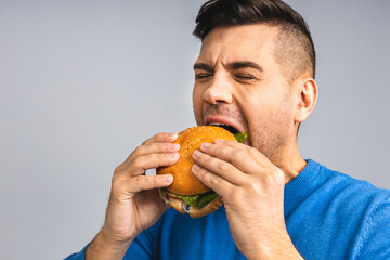 Young ukrainian man holding a piece of hamburger. Student eats fast food. Burger is not helpful food. Very hungry guy. Diet concept. Isolated over grey white background.