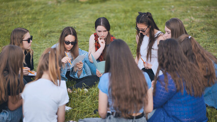 A group of female students are sitting in a circle on a meadow for collective work with notebooks.