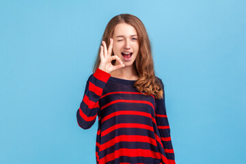 Portrait of woman wearing striped casual style sweater standing showing ok sign and winking to camera, approval sign, agreeing with suggestion. Indoor studio shot isolated on blue background.