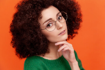 Closeup portrait of pleasant looking woman with Afro hairstyle wearing green casual style sweater and eyeglasses, keeping hand under chin. Indoor studio shot isolated on orange background.