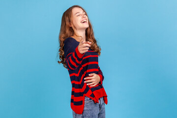 Lol, hey you! Excited joyful woman wearing striped sweater laughing out loud, holding her belly and pointing finger to camera, making fun of people. Indoor studio shot isolated on blue background.