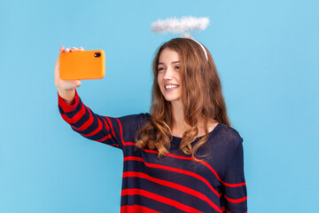 Woman blogger wearing striped casual style sweater and nimb over head, holding phone, taking selfie or having livestream, taking with followers. Indoor studio shot isolated on blue background.