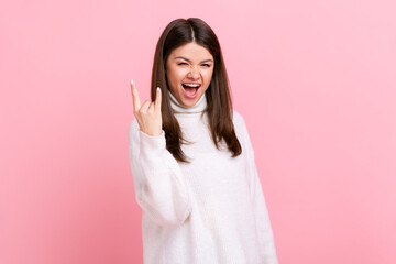 Young adult excited female with dark hair showing rock and roll gesture, yelling with excitement, wearing white casual style sweater. Indoor studio shot isolated on pink background.
