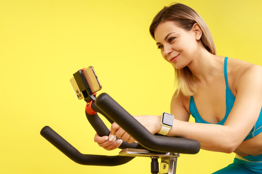 Closeup of athletic woman riding exercise bike and using her mobile phone for broadcasting her cardio workout online, wearing blue sportswear. Indoor studio shot isolated on yellow background.