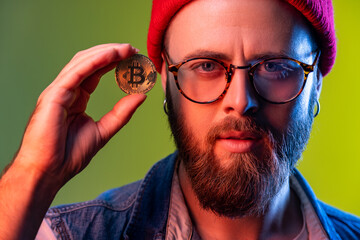 Serious hipster man holding in fingers and showing golden coin bitcoin crypto currency, electronic virtual money, wearing beanie hat. Indoor studio shot isolated on colorful neon light background.