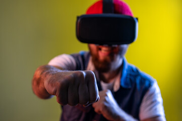 Hipster man in virtual reality glasses on head playing fighting game, holding clenched fists up to camera, ready to boxing. Indoor studio shot isolated on colorful neon light background.