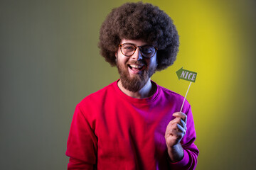 Happy positive hipster man with Afro hairstyle having event, holding party props with nice word in hands, wearing red sweatshirt. Indoor studio shot isolated on colorful neon light background.
