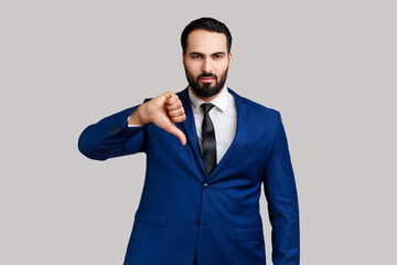 Bearded man criticizing bad quality with thumbs down displeased grimace, showing dislike gesture, expressing disapproval , wearing official style suit. Indoor studio shot isolated on gray background. - obrazy, fototapety, plakaty