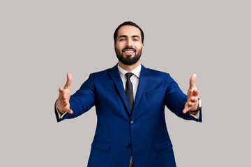 Positive bearded man giving free hugs with outstretched hands, welcoming inviting to embrace, support and care, wearing official style suit. Indoor studio shot isolated on gray background.