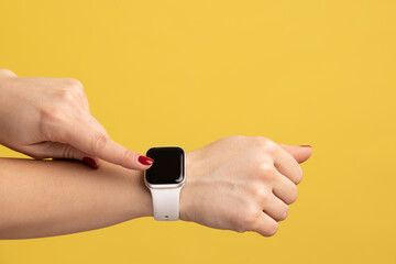Closeup side view of woman hand with smartwatch touching touchscreen, arm with wristtwatch with white strap, technology device. Indoor studio shot isolated on yellow background.