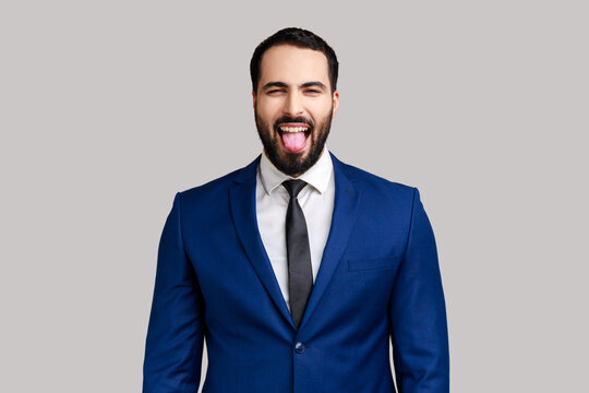 Portrait of playful bearded businessman showing tongue, having fun, fooling around, childish manners, wearing official style suit. Indoor studio shot isolated on gray background.