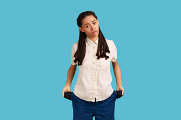 Unemployment and bankruptcy. Upset poor woman with dreadlocks turning out empty pockets, worried about debts, no cash for living, wearing white shirt. Indoor studio shot isolated on blue background.