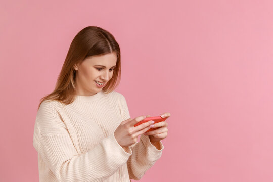 Portrait of happy positive blond woman holding smart phone in hands and playing video games, successfully completing level, wearing white sweater. Indoor studio shot isolated on pink background.