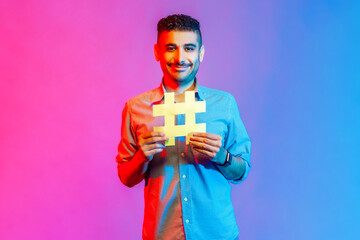 Portrait of man in shirt with toothy smile on his face holding hashtag symbol, recommending popular topics, internet trends. Indoor studio shot isolated on colorful neon light background.