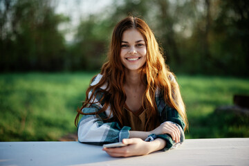 Girl with phone in hand sitting in nature in the park at the table smiling beautifully and looking at the camera with her red hair lit by the sunset sunlight of summer