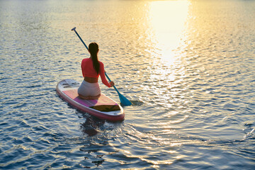 Back view of healthy woman with slender body sitting on sup board and using long paddle for rowing. Caucasian brunette in sportswear doing water activity during evening time outdoors.