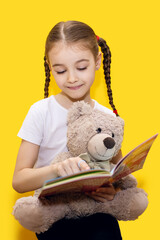A girl is reading a book and holding a teddy bear on a yellow background. A cute girl with pigtails...