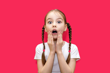 A girl with pigtails on a red background with a surprised look holds her head. Shocked little girl...