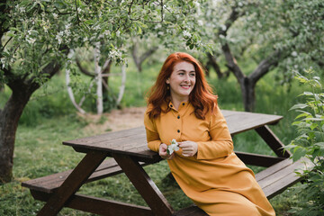 Naklejka premium Portrait of gorgeous middle aged woman in casual dress relaxing in a blooming spring garden. Happy senior woman smiling and looking at camera. Red-haired mature lady posing outdoors in a spring day.