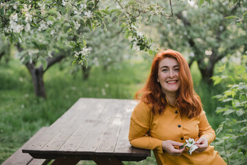 Portrait of gorgeous middle aged woman in casual dress relaxing in a blooming spring garden. Happy senior woman smiling and looking at camera. Red-haired mature lady posing outdoors in a spring day.