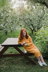 Naklejka premium Portrait of gorgeous middle aged woman in casual dress relaxing in a blooming spring garden. Happy senior woman smiling and looking at camera. Red-haired mature lady posing outdoors in a spring day.