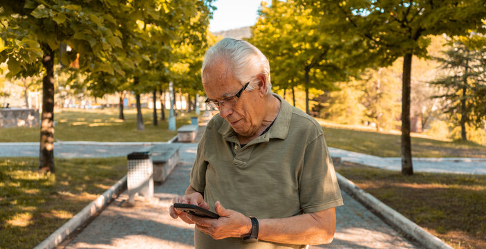 Hombre mayor en el parque, mirando su tel&eacute;fono m&oacute;vil con expresi&oacute;n de preocupaci&oacute;n. Fotograf&iacute;a horizontal con espacio para texto.