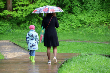 Summer rain, woman with kid daughter walking with umbrella in green park