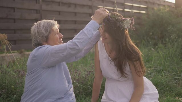 Retired mother arrange flower crown on daughter head, beautiful nature, summer garden, smiling faces, warm sun light, pure and eternal love concept