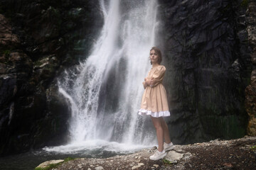 Lonely girl in a short beige dress stands next to the waterfall.