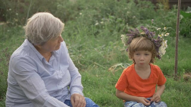 Portrait of old woman with gray hair and blond child with flower crown sit on green grass