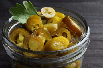 Glass jar with slices of pickled green jalapeno peppers on black wooden table, closeup