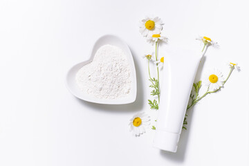 White plastic tube with cream, balm or lotion and mineral cosmetic clay in ceramic bowl with fresh chamomile flowers on white background top view. Natural herbal cosmetic concept.